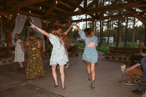 Reception guests dance playfully in a line against a wooded outdoor setting with string lights overhead.