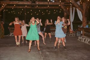 Wedding guests dance together in colorful dresses under a wooden pavilion at dusk.