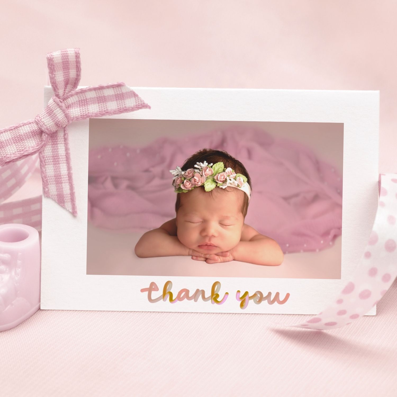 Pink thank you card featuring a sleeping newborn with floral headband on soft pink background with gingham bow decoration.