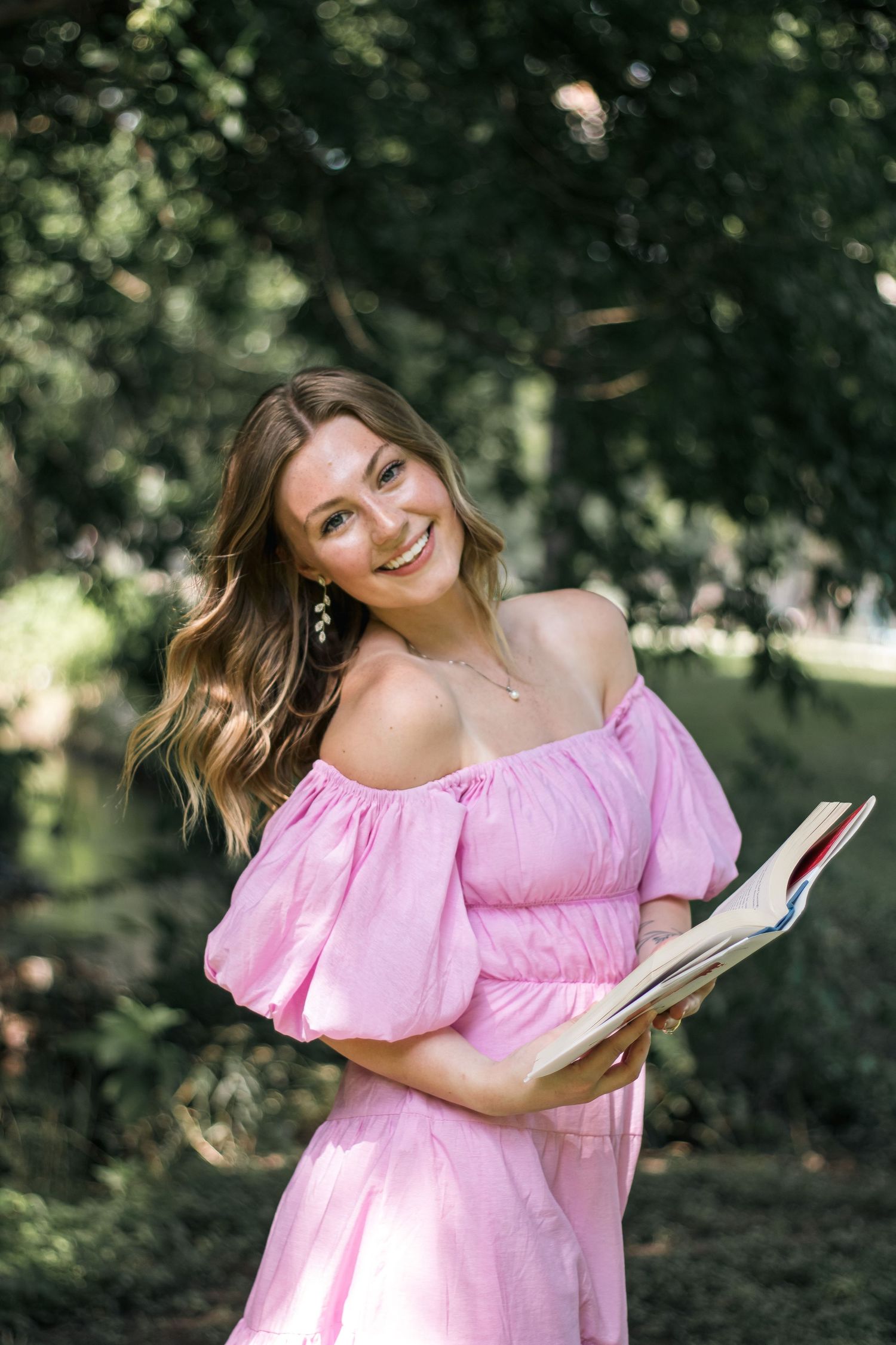 Smiling young woman in pink off-shoulder dress holding an open book outdoors in a lush green park.