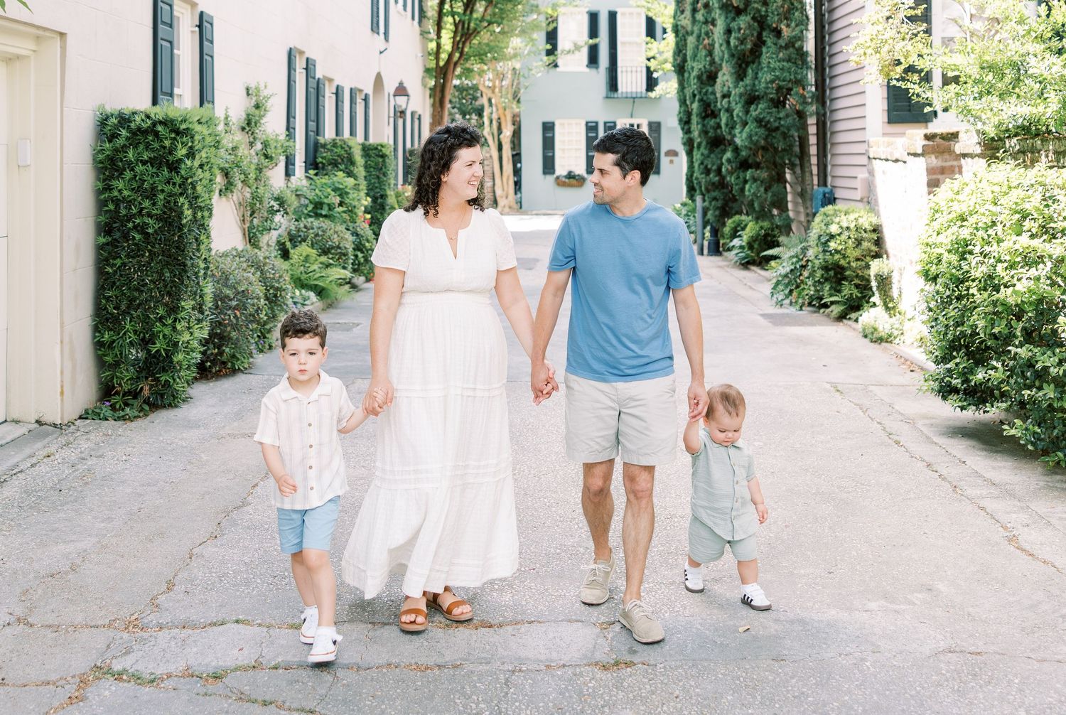 Family walks together along charming tree-lined alley with white building and greenery in summer.