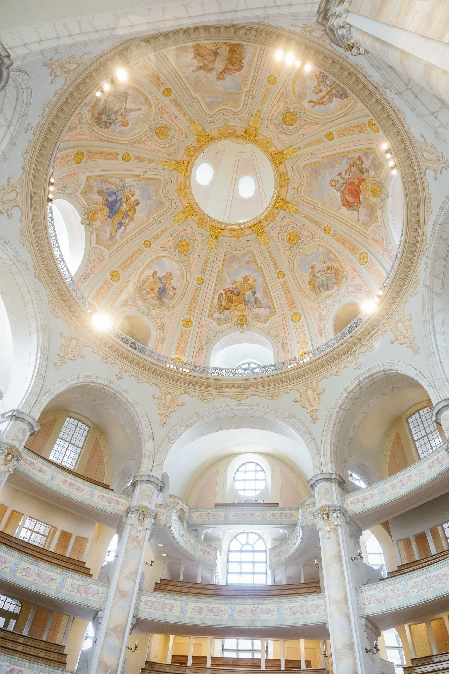 Ornate baroque church interior with white and gold decorated domes, classical columns and arched windows illuminated by natural light.