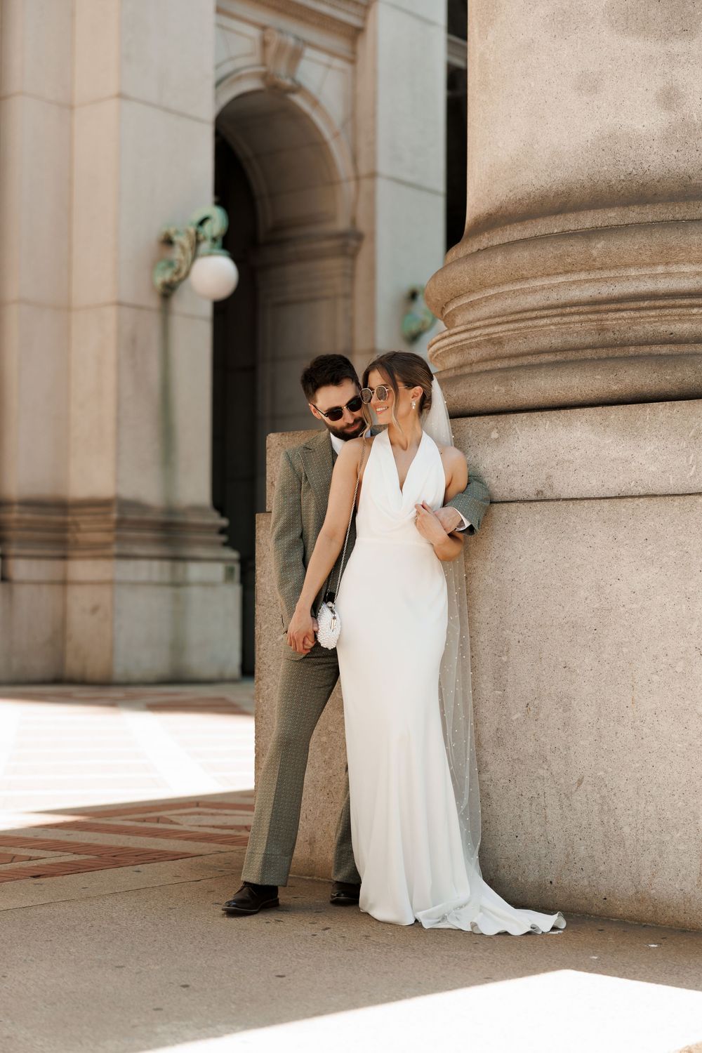 Bride in flowing white gown and groom share an intimate moment leaning against a stone column.