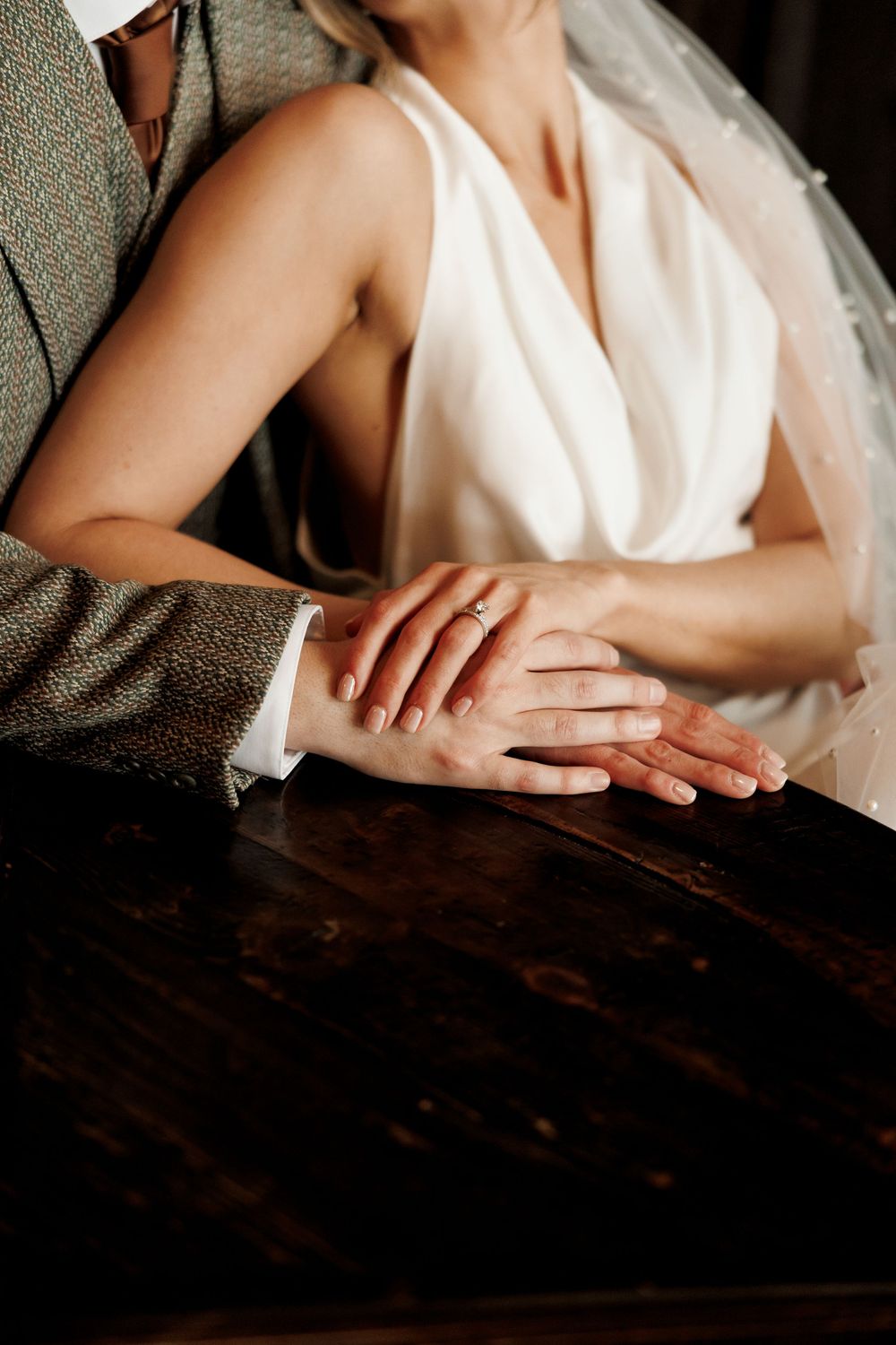 Bride and groom holding hands on dark wooden table, wedding ring visible, bride in white halter gown.