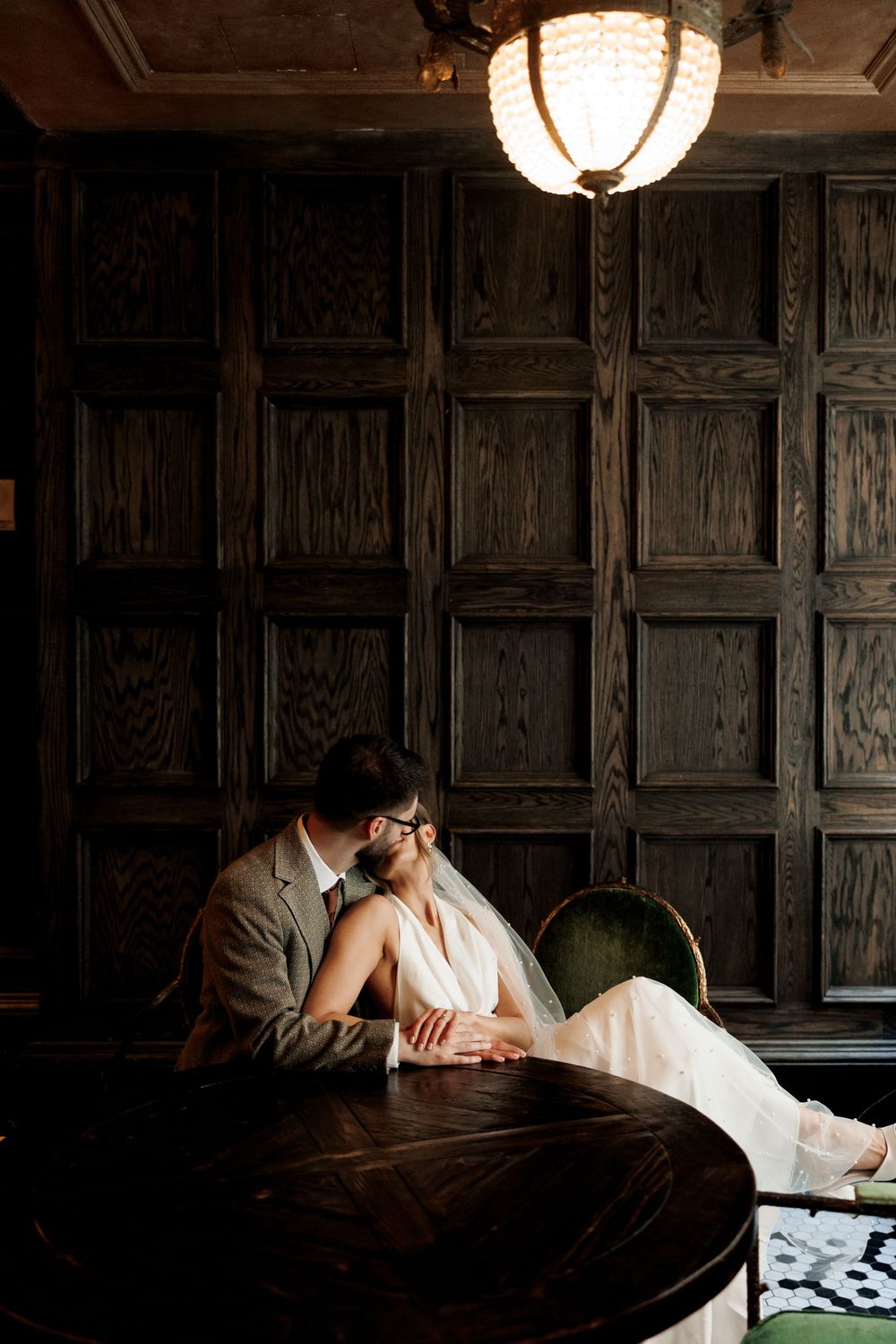 Bride and groom sitting intimately at dark wooden table in elegant paneled room with vintage chandelier.