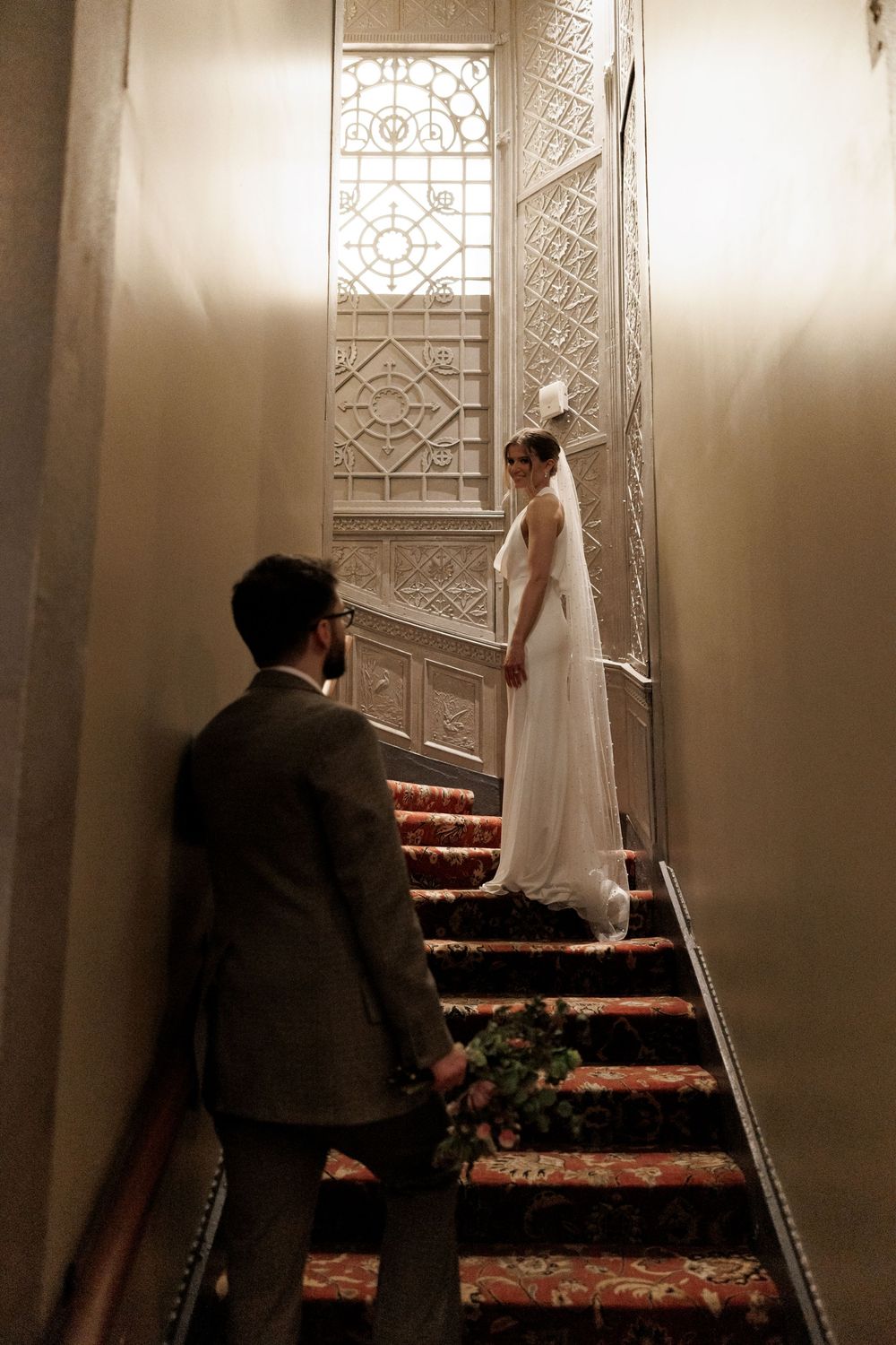 Groom holding bouquet watches bride in white gown and veil on ornate staircase with stained glass window.