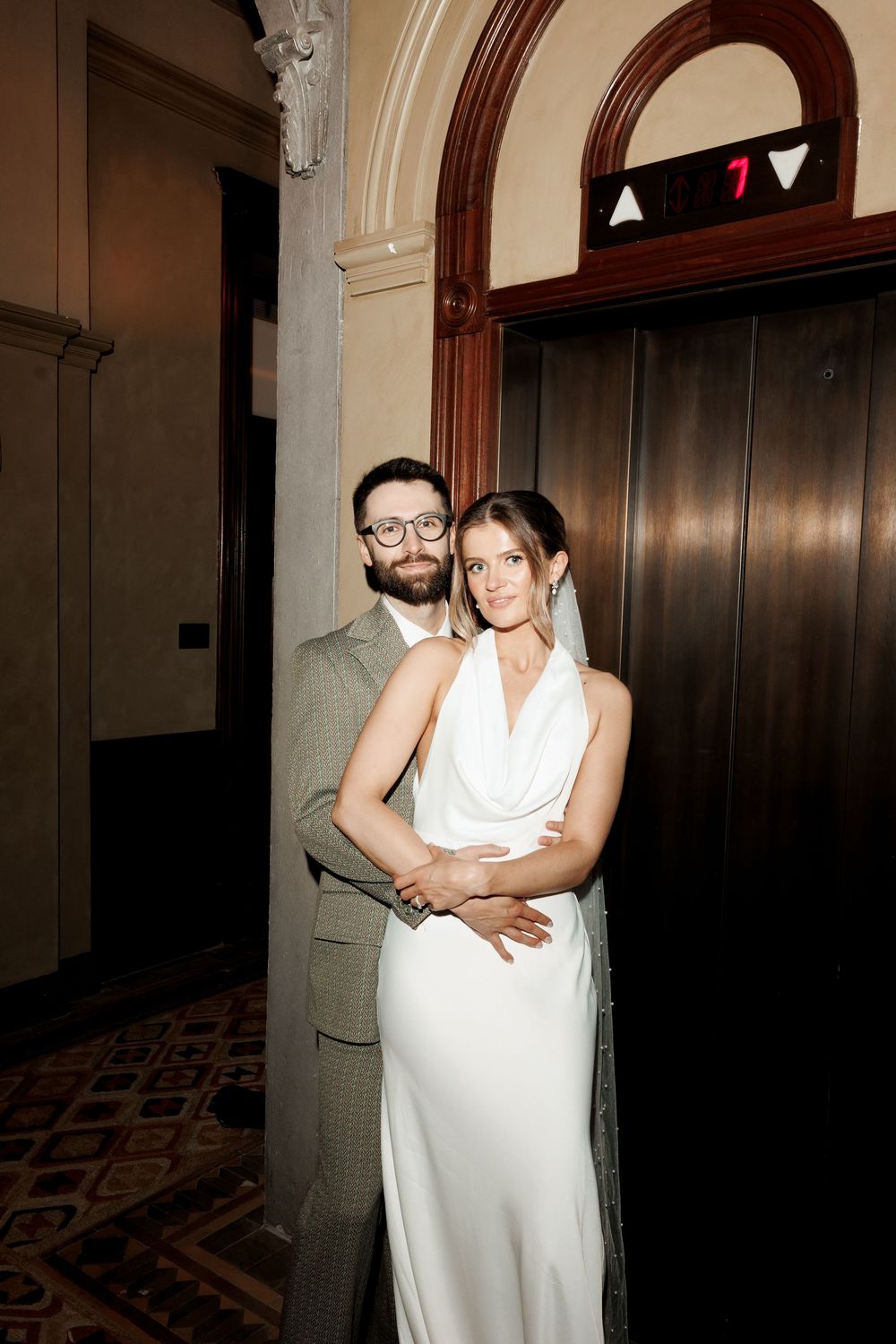 Bride in white cowl-neck gown and groom in plaid suit posing by vintage elevator on floor seven.