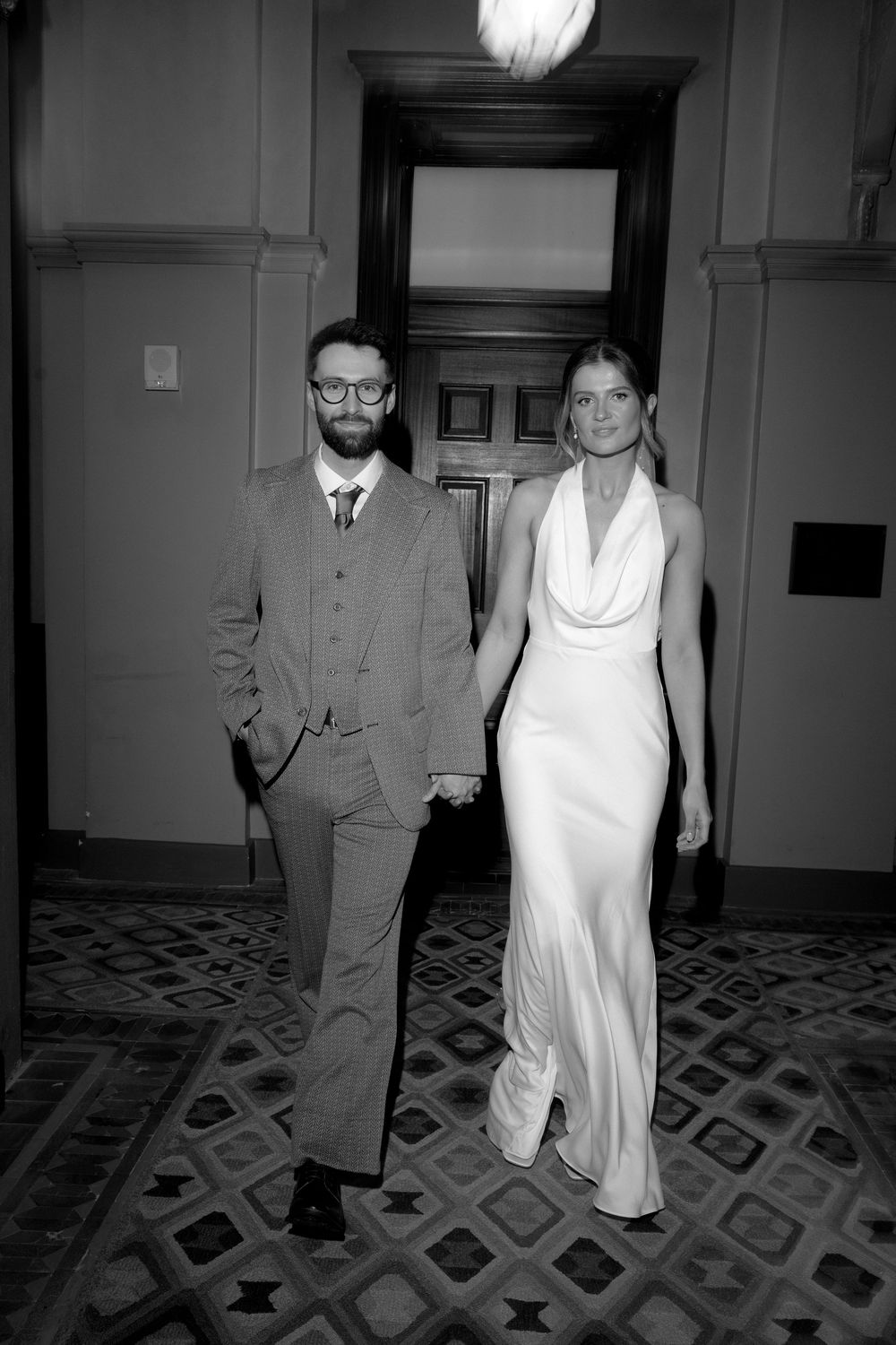 Black and white photo of bride in sleek white gown and groom in suit walking hand in hand.