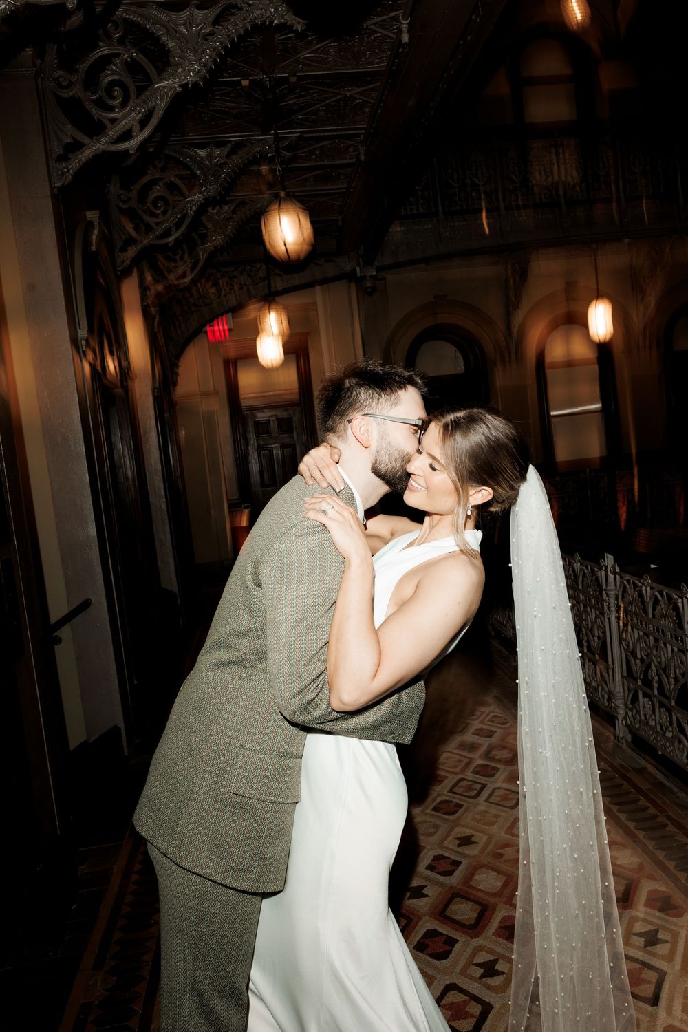 Bride and groom embracing tenderly in a beautifully lit historic venue with intricate ceiling details.