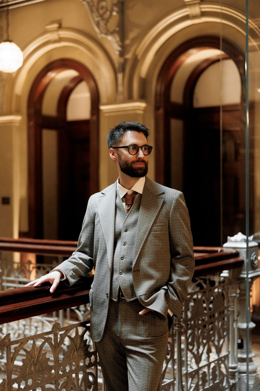 Dapper groom in gray three-piece suit and glasses stands confidently by decorative iron railing in elegant venue.