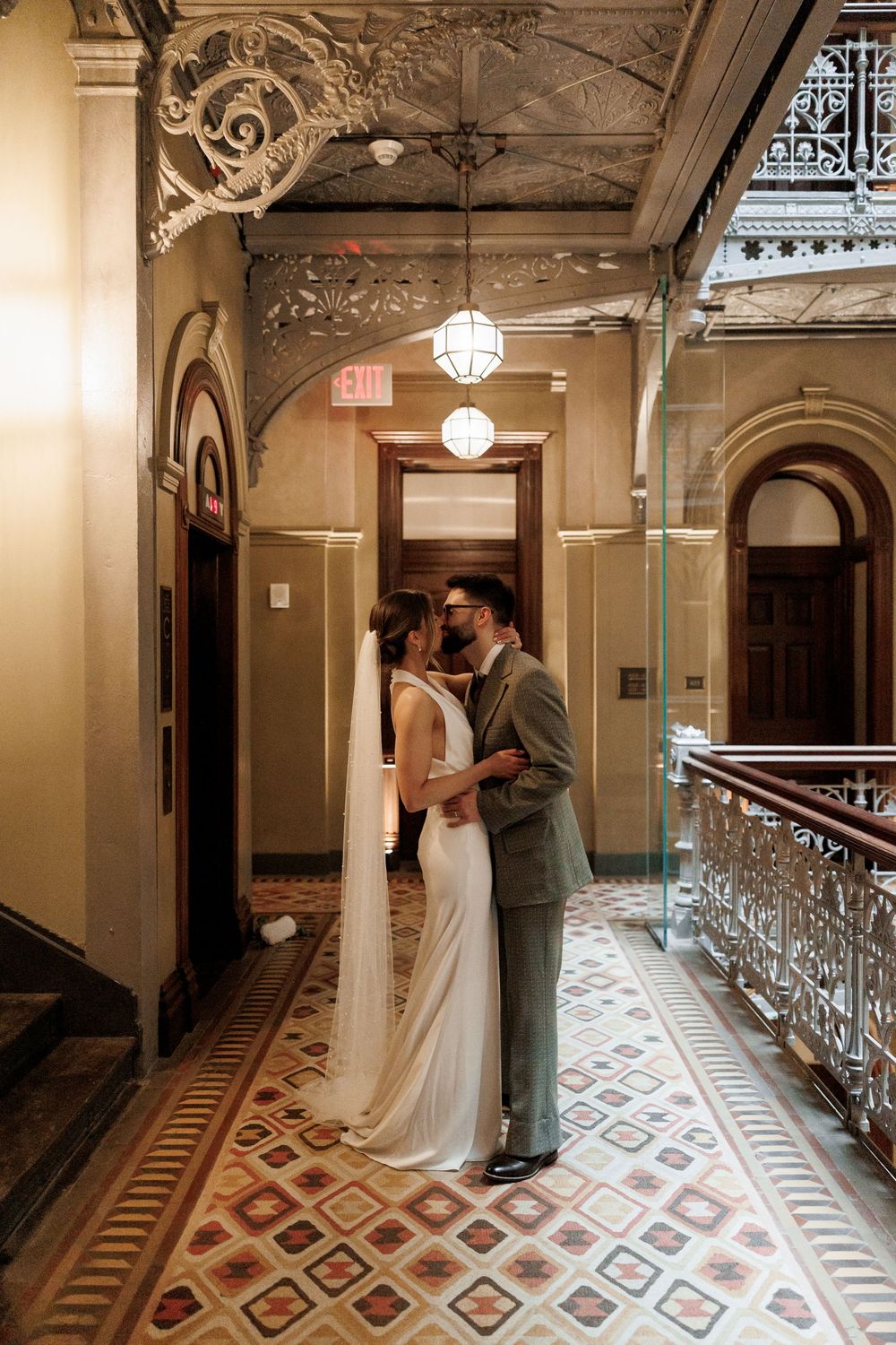 Bride and groom share intimate moment in sunlit ornate hallway with decorative tiled floors and ironwork.