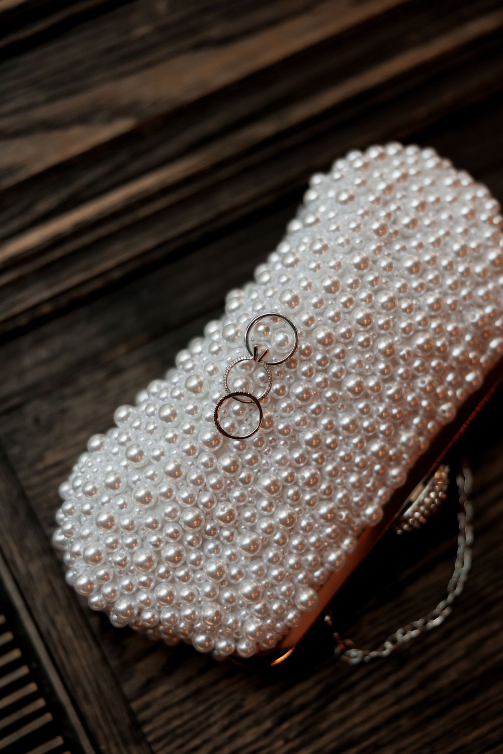 Two gold wedding rings rest elegantly on a pearl-covered bridal clutch purse on wooden surface.