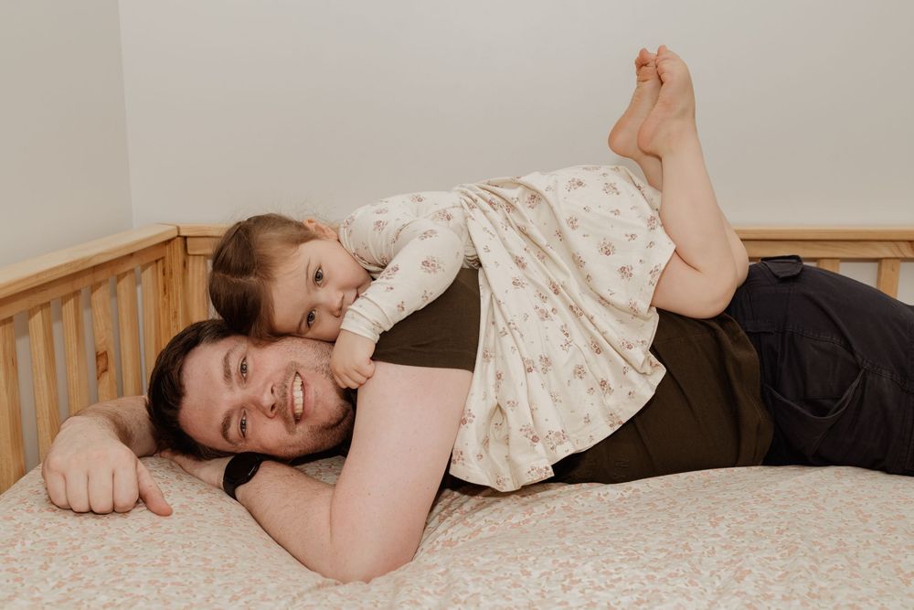 A father lying down on his daughter's bed on his stomach. His daughter is on his back, they are smiling and giggling at the camera.