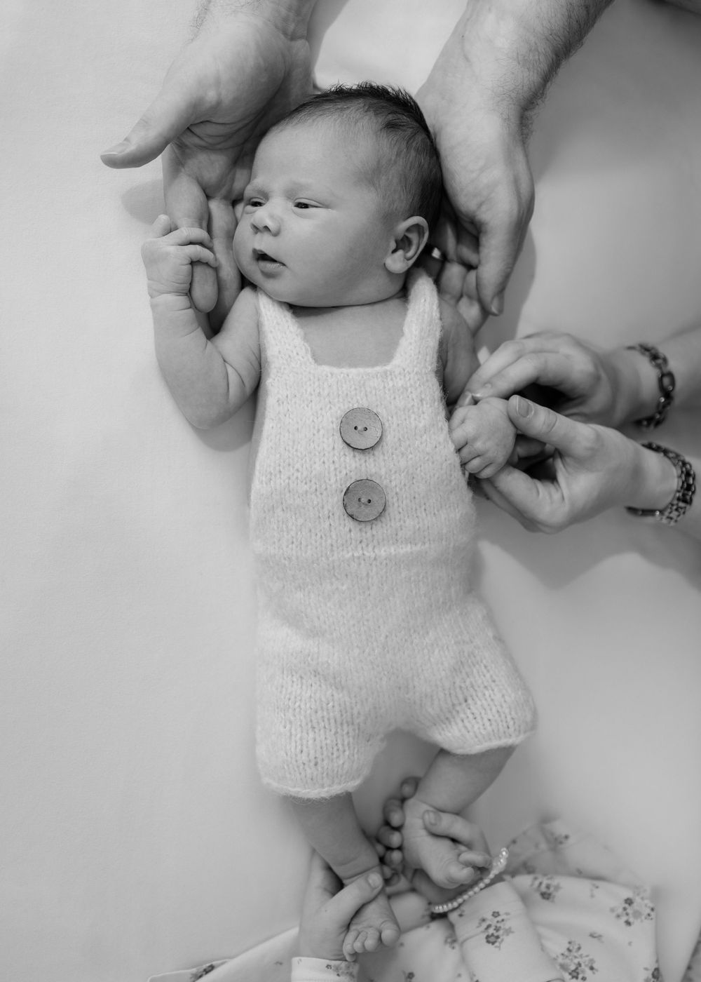 Black & white of newborn stretched out on bed. Big sister holding baby's feet. Mum holding baby's hand/arm. Dad holds baby's head