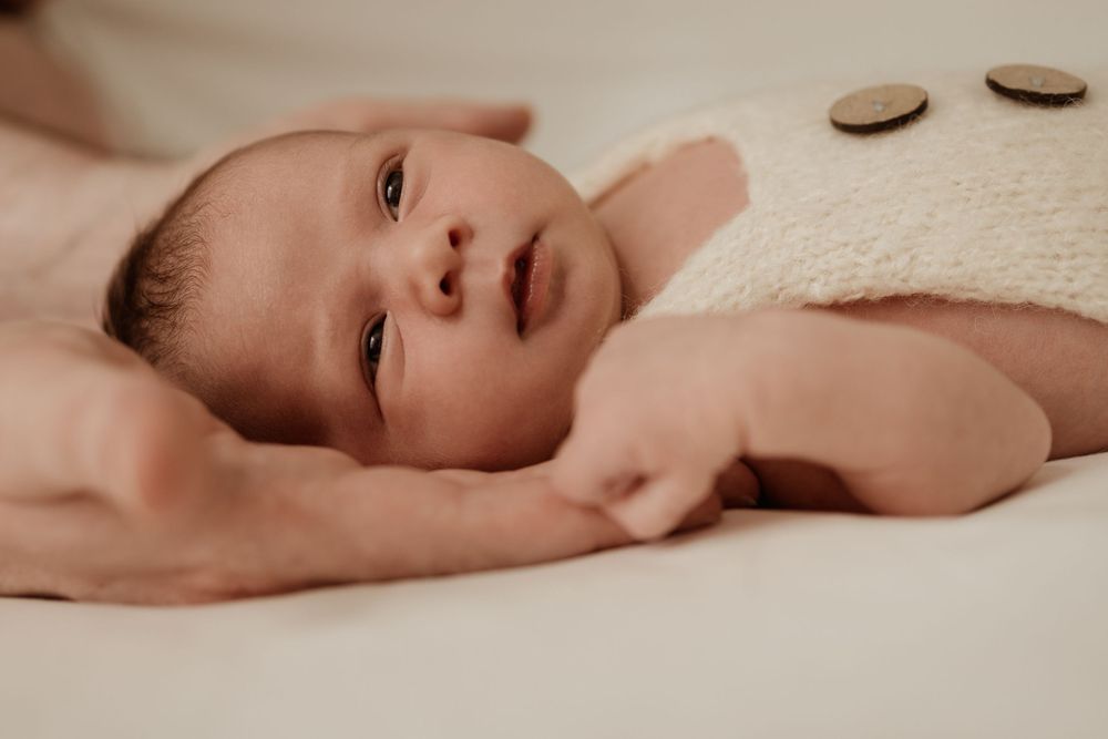 close up of newborn face. Glassy eyes staring into distance. Dad's hands cradling baby's head. Cream knitted buttoned overalls