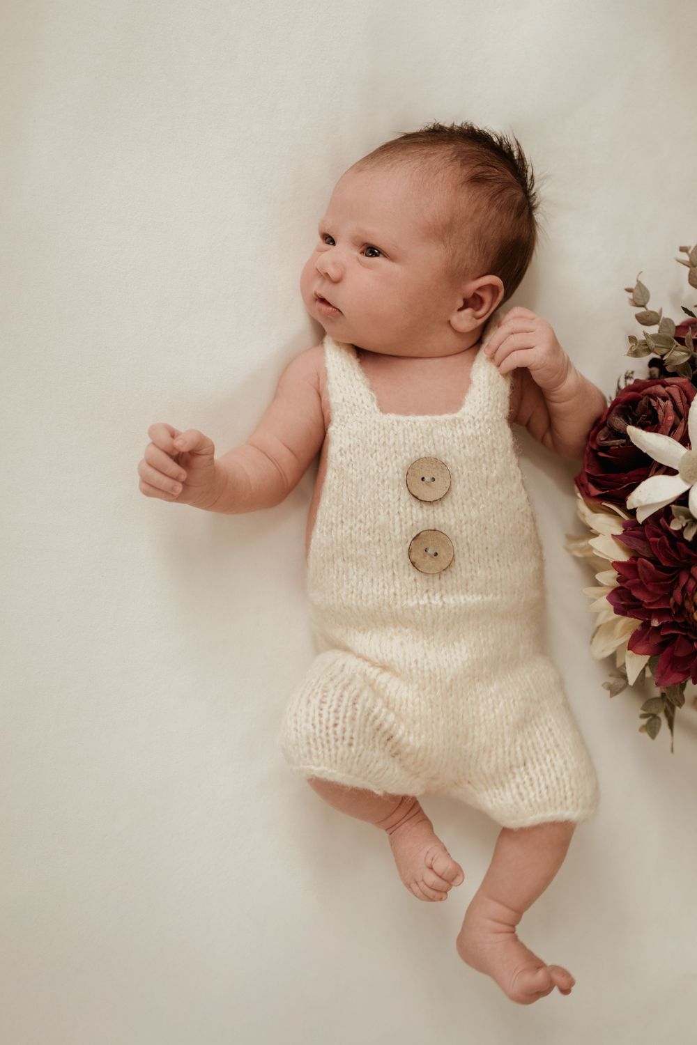 Newborn lying on the bed wearing knitted jumpsuit with big wooden buttons. Her parents wedding bouquet off to the side.