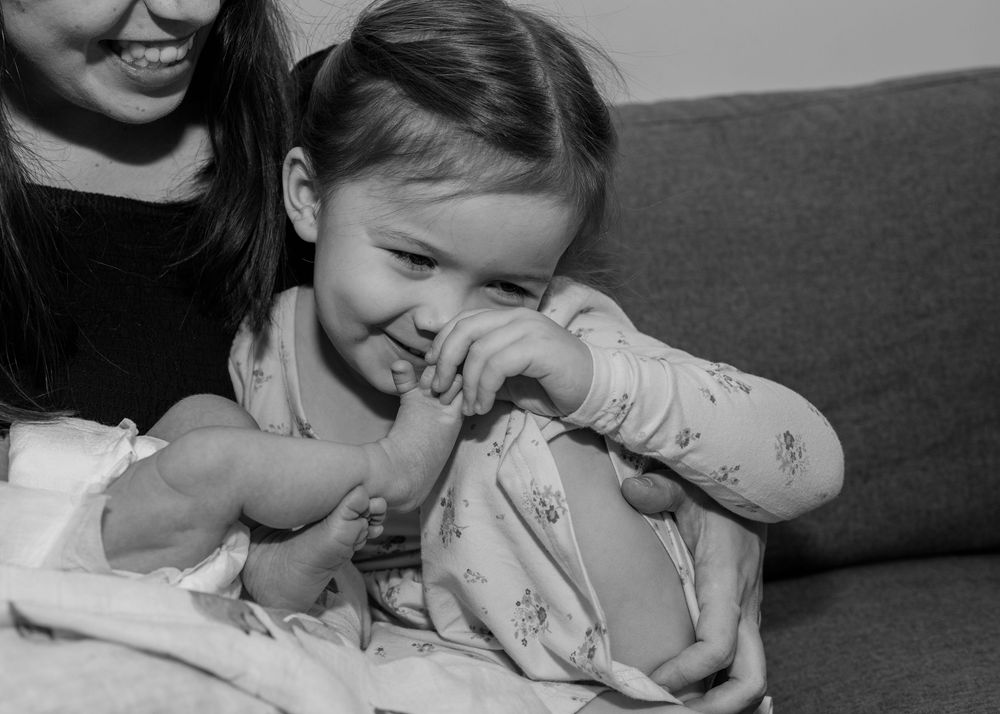 Big sister sitting on the couch with her mother as she playfully tickles her newborn sister's toes. Close up of details. Black and white