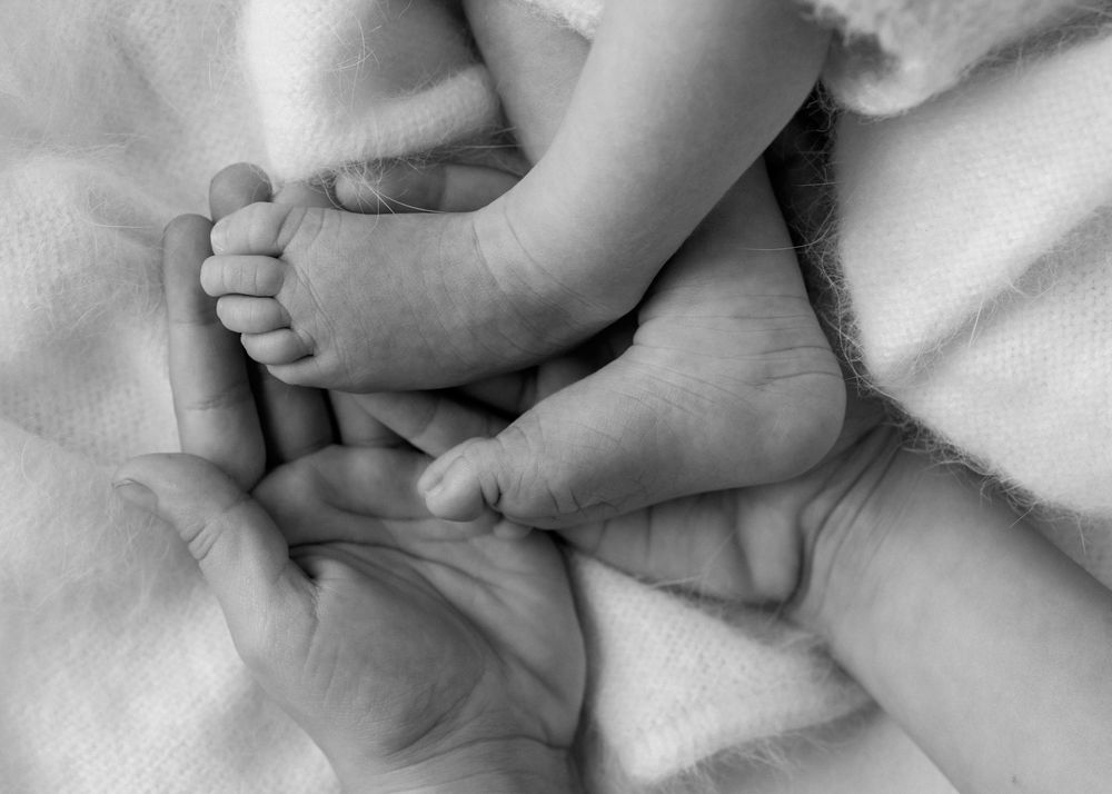 Close up macro Black and white image of big sister holding her newborn baby sister's feet as she lays on a wooden blanket
