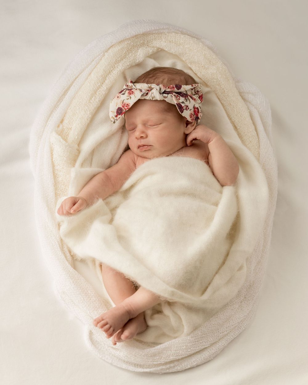 Newborn baby sleeping - wrapped in white wooden blanket, with textured knits. Floral headband, legs crossed. Newborn photography.