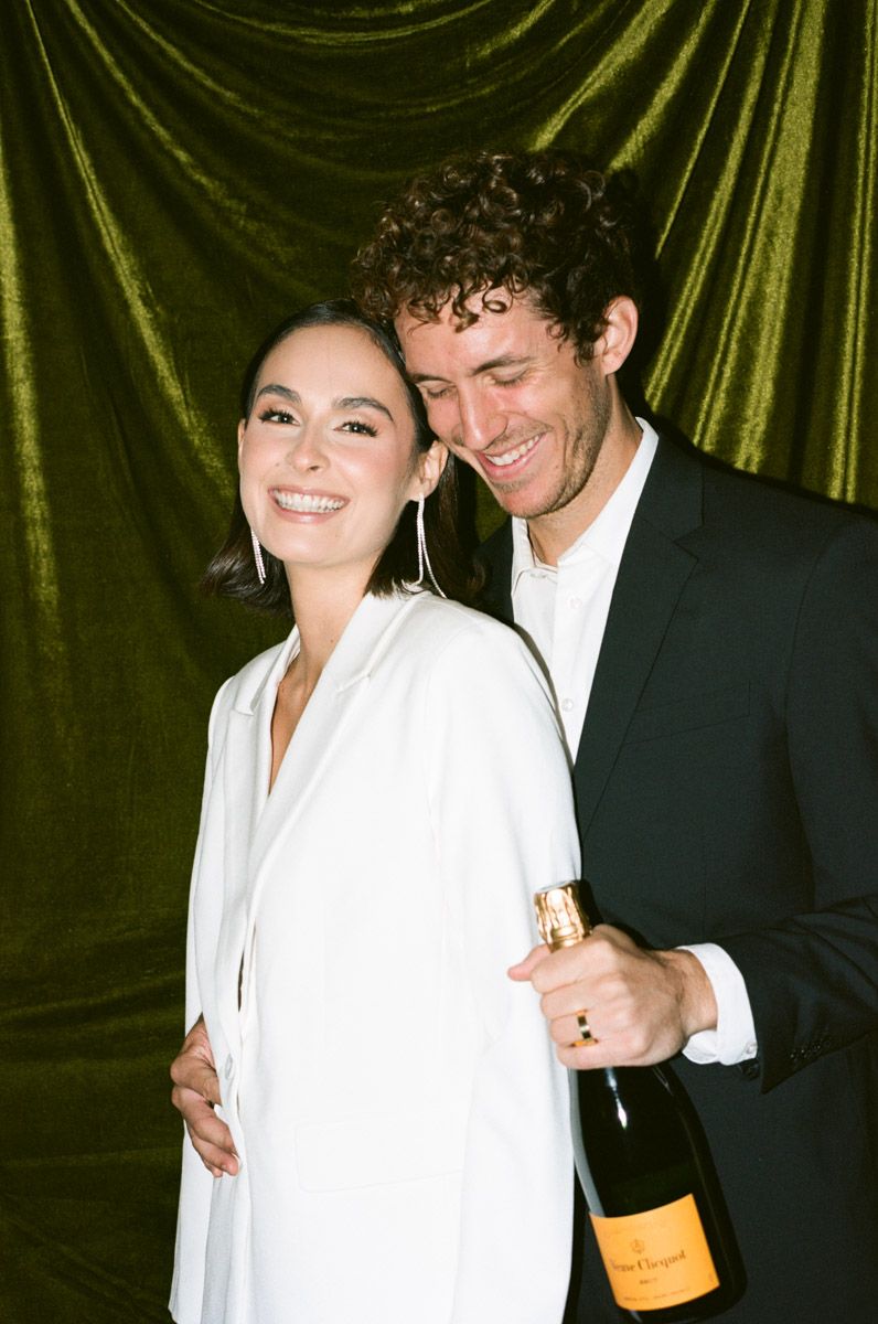 Couple celebrating with Veuve Clicquot champagne bottle, woman in white blazer and man in black suit, green velvet backdrop.