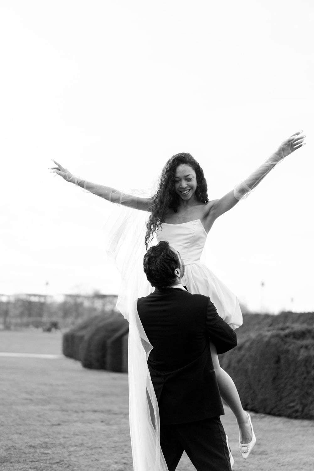 Black and white photo of couple in wedding attire outdoors, with bride lifted in celebratory pose with arms outstretched.