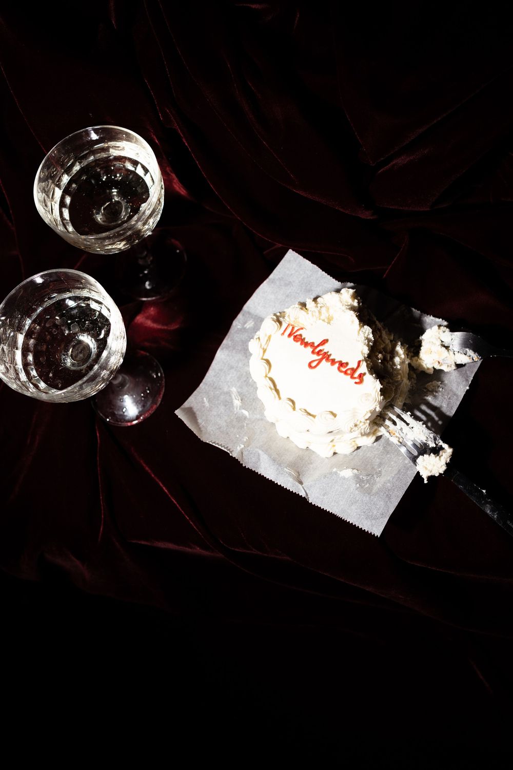 Two glasses of clear wine and a small white frosted cake with 'Happy Birthday' text on dark velvet background.