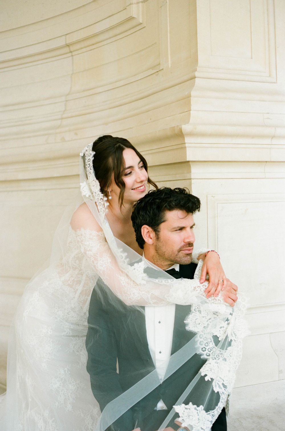 Elegant wedding portrait showing romantic embrace with delicate lace veil against white architectural backdrop.