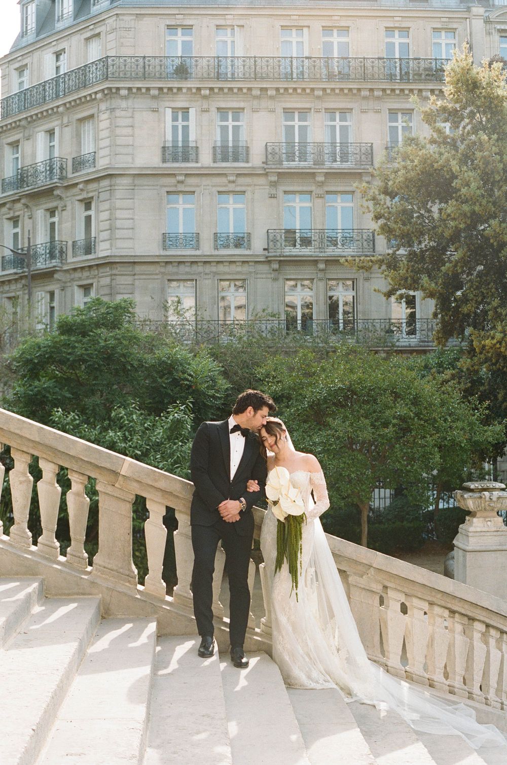 A couple in wedding attire poses on an elegant stone balcony with classical European architecture in the background.