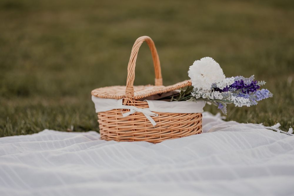 Wicker picnic basket with white bow and wildflowers on a white blanket in a green field.
