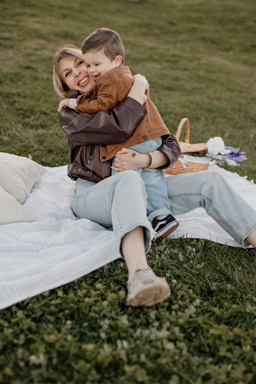 A woman and young boy share a warm hug on a white blanket in a green park during a picnic.