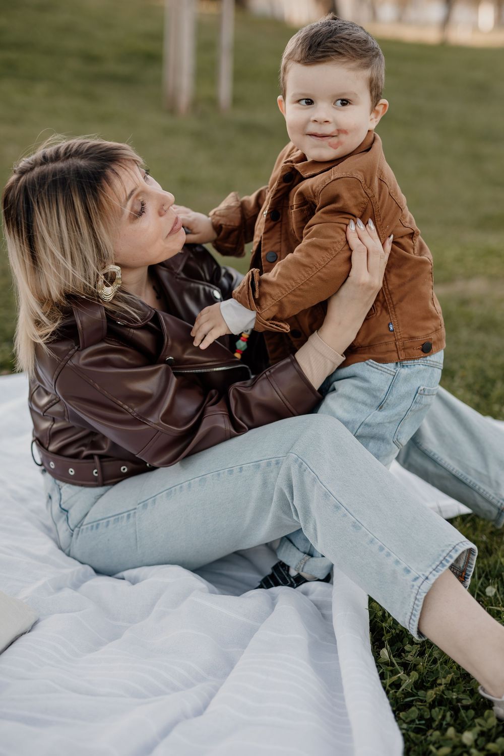A mother in a brown leather jacket sits on a blanket outdoors, lovingly holding her smiling toddler son.