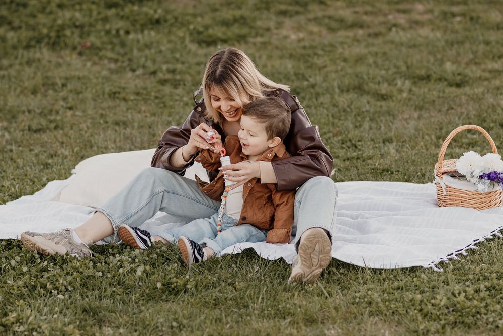 Mother and child sharing a joyful picnic moment on a white blanket in a green park with a basket nearby.