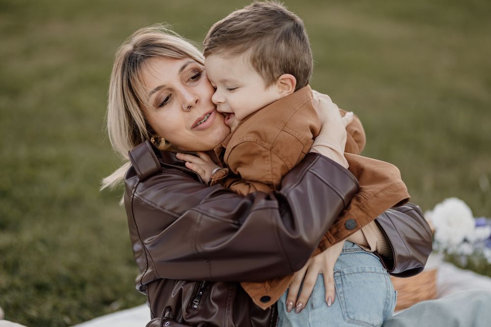 A mother in a brown leather jacket hugs her young son outdoors on a green lawn during a family photo session.