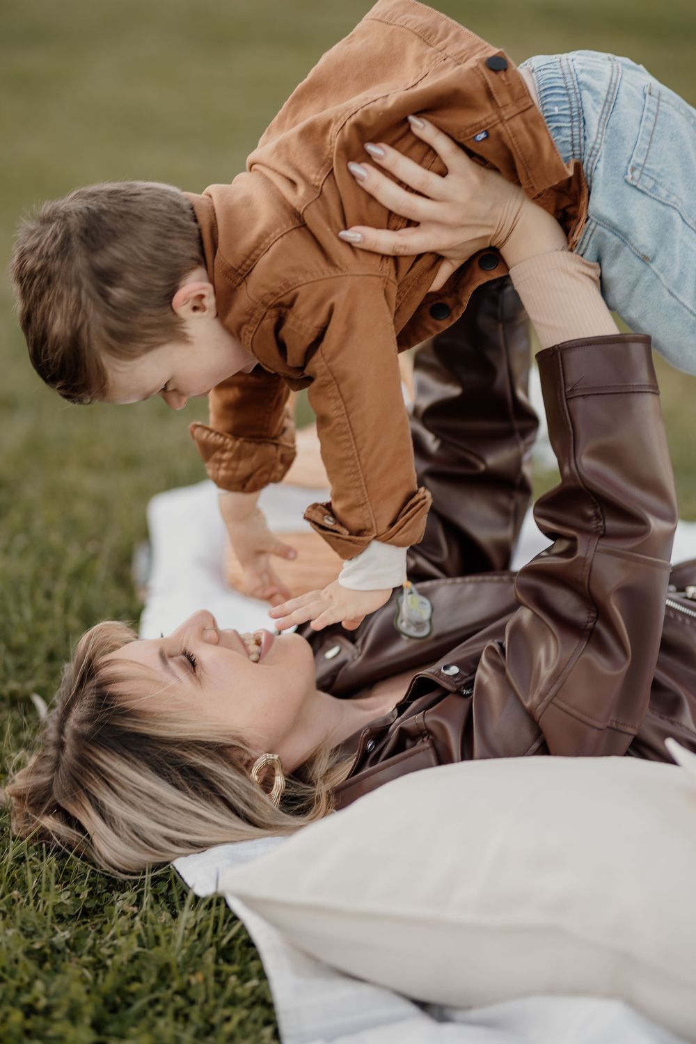 A mother lies on grass, playfully lifting her young son above her, both smiling and enjoying a fun outdoor moment.