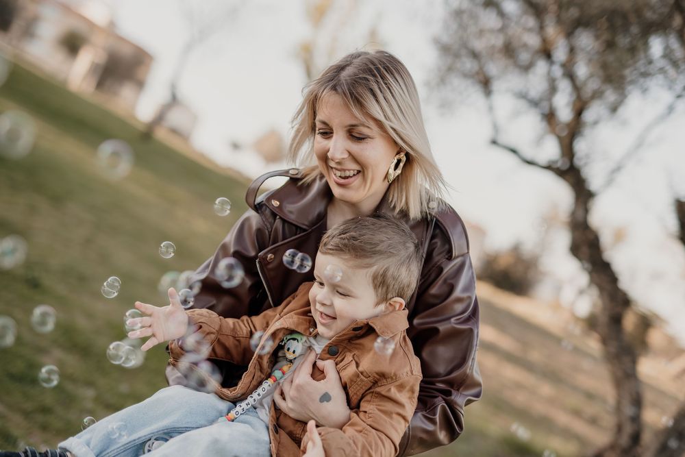 A smiling woman and toddler in brown jackets play with bubbles outdoors on a sunny day.