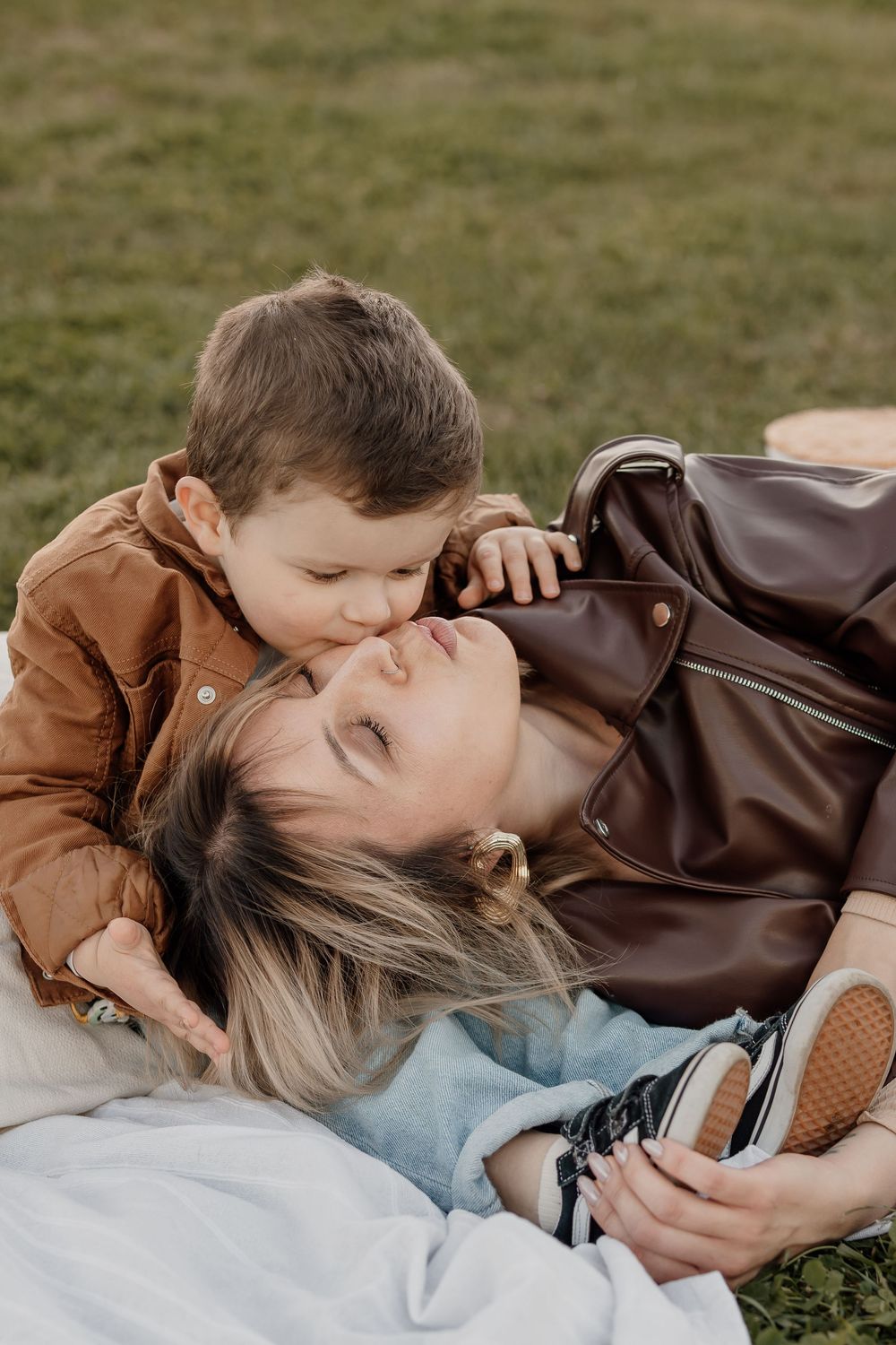 A young boy tenderly kisses his mother's cheek as she lies on grass during an outdoor family moment.
