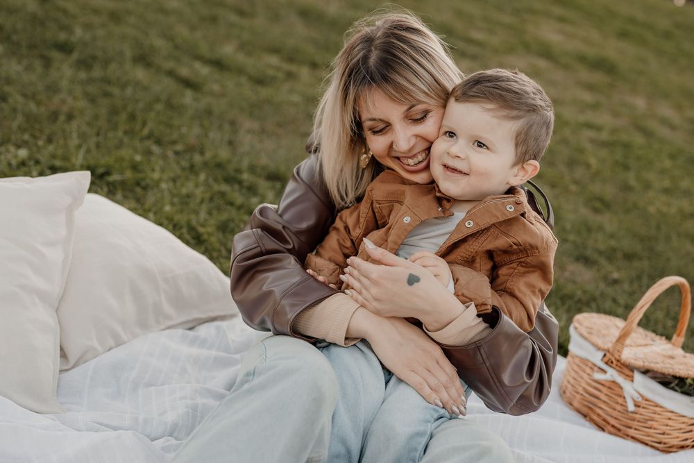 A smiling mother hugs her toddler son on a white blanket during an outdoor picnic on green grass.