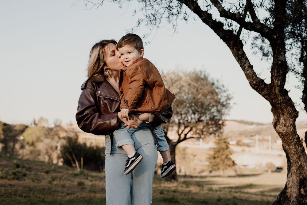 A mother in a brown leather jacket holds her toddler son outdoors near a tree in a scenic countryside setting.