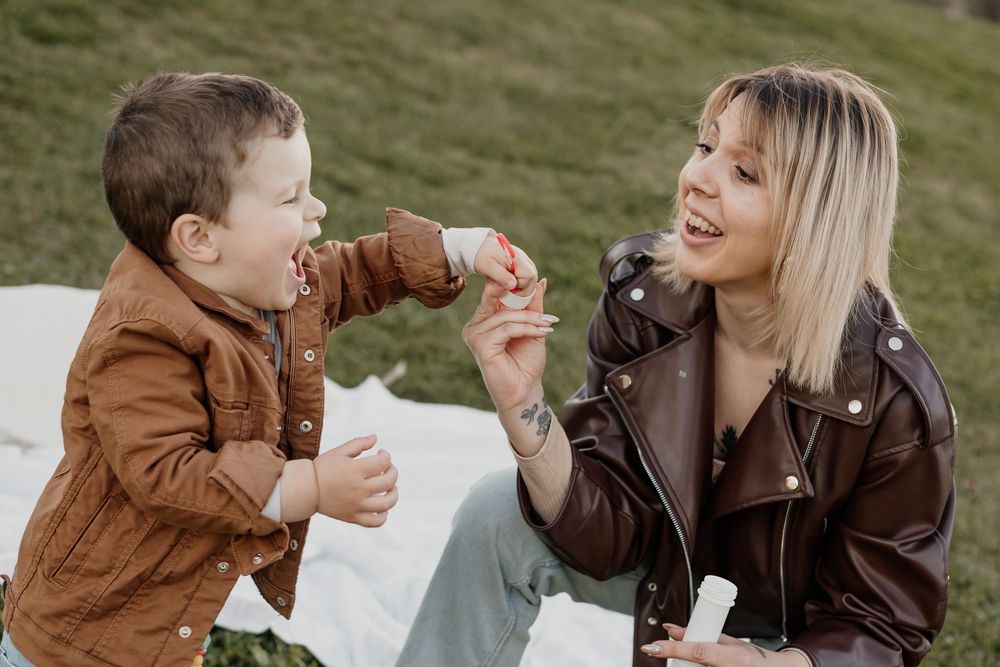 A smiling woman in a brown leather jacket blows bubbles with a laughing toddler boy outdoors on a sunny day.