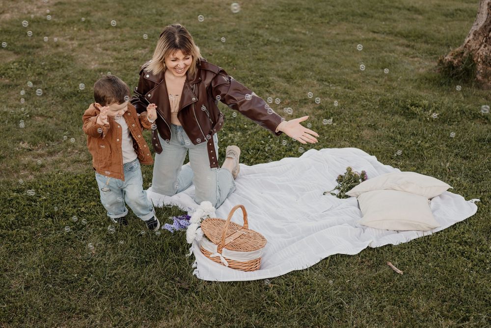 Woman and child playing with bubbles on a white blanket during a sunny outdoor picnic in a green park.