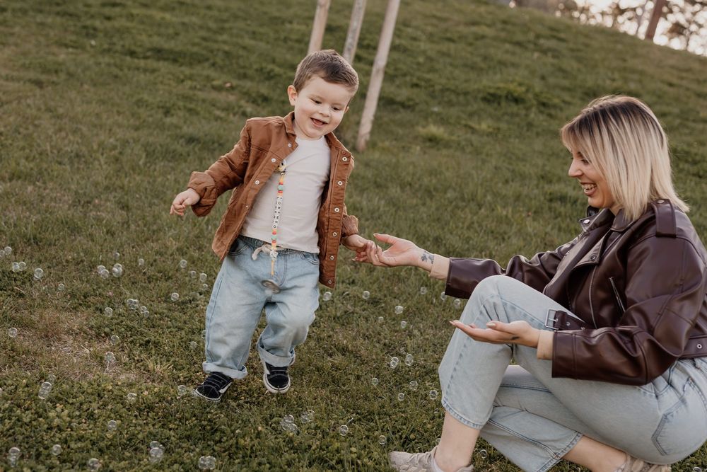 A young toddler boy plays outdoors on green grass while a smiling blonde woman reaches out her hand to him.