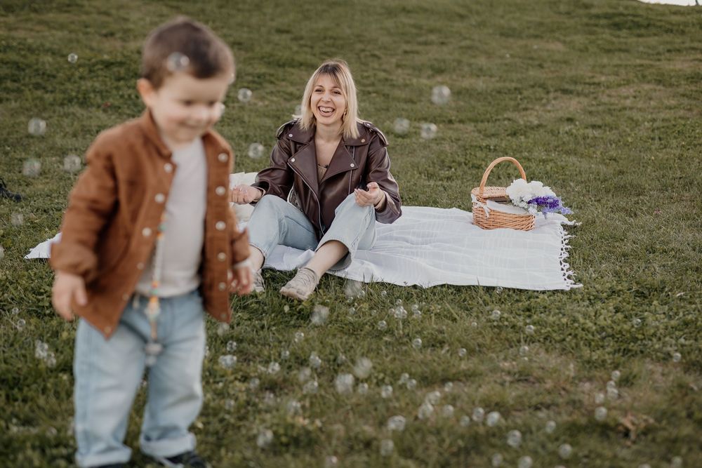 A mother sits on a blanket in a park while her young son plays with bubbles during a sunny picnic.