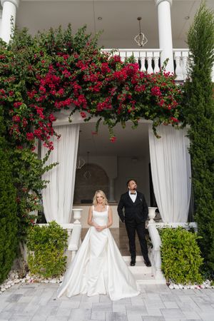 Bride and groom pose under vibrant red bougainvillea at elegant white villa entrance with draped curtains.