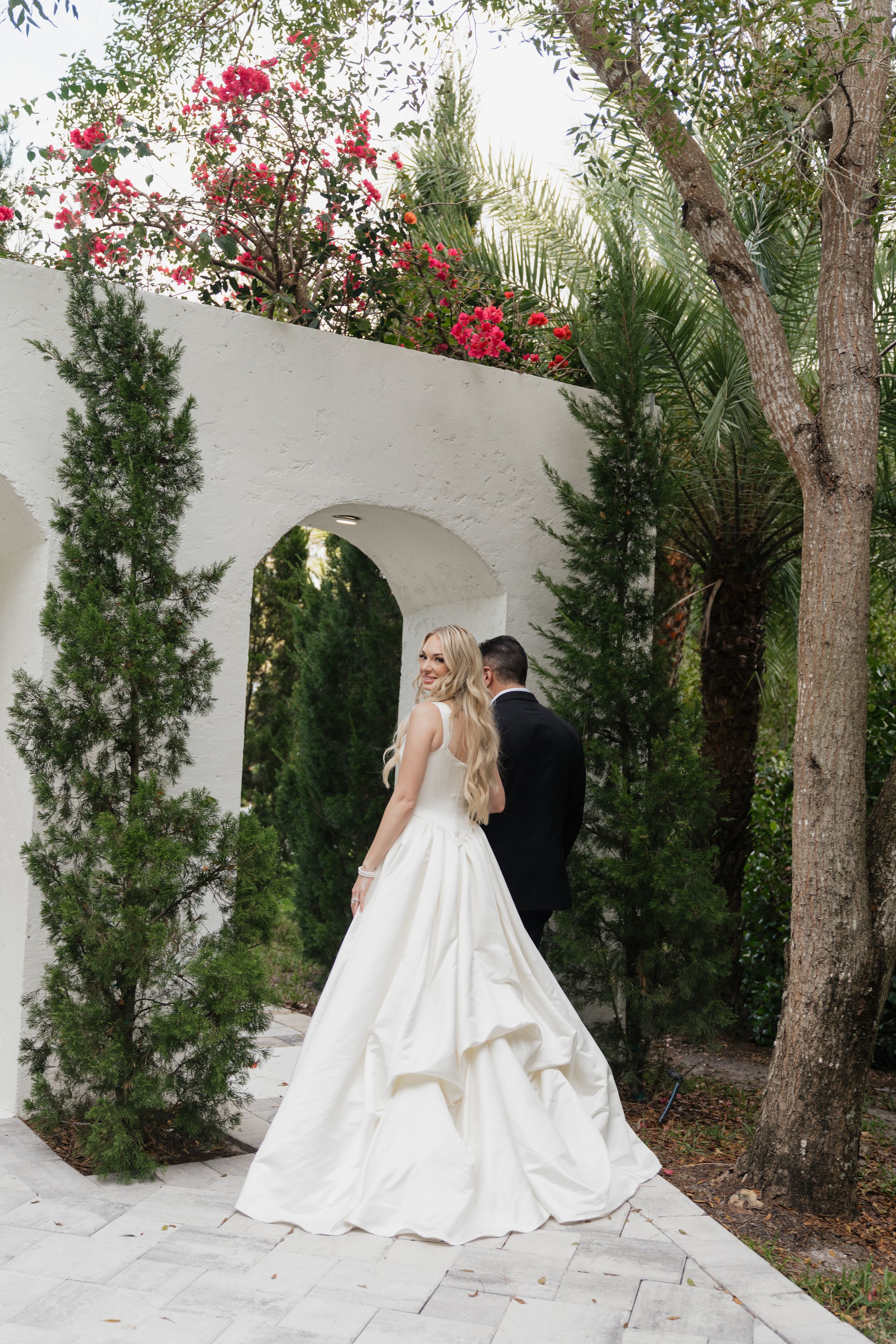 Bride in white ball gown and groom in black tuxedo walk through a white archway surrounded by lush greenery and red flowers.