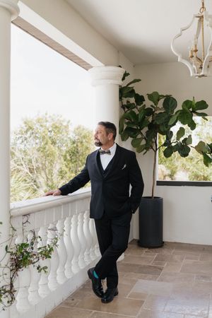 Groom in black tuxedo with bow tie stands on elegant balcony beside fiddle leaf fig tree, gazing into distance.