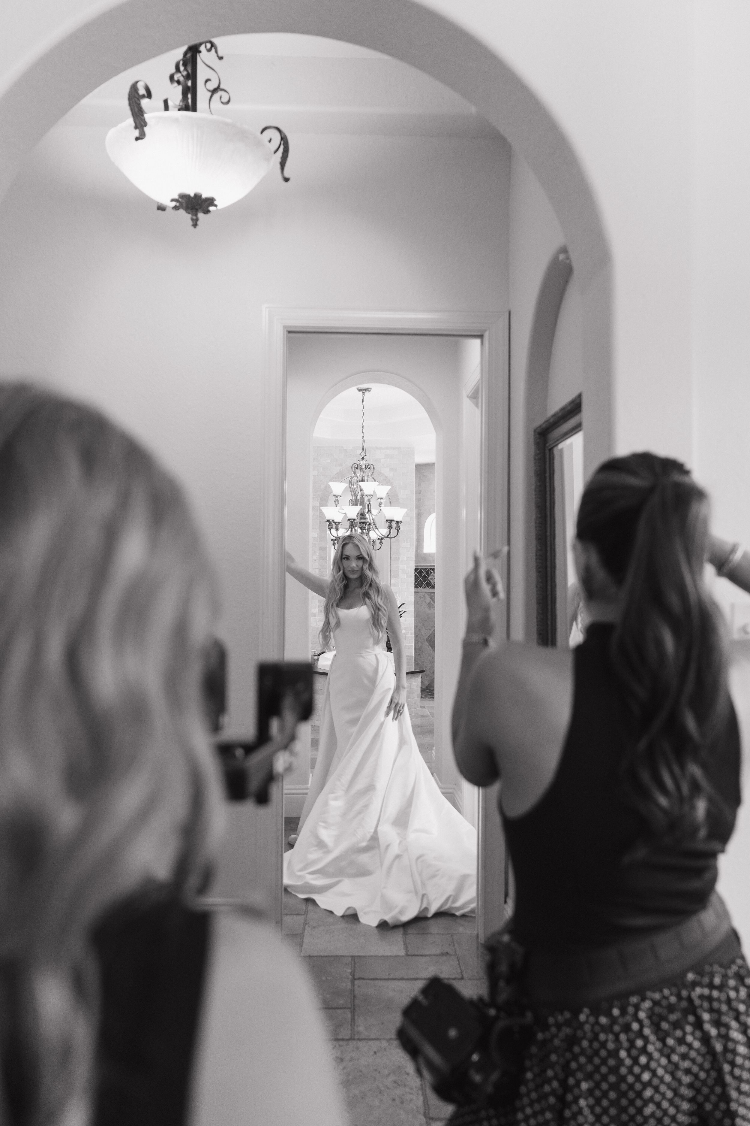 Black and white photo of a bride in a white gown posing in a doorway as bridesmaids photograph her.