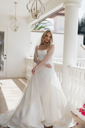 Elegant bride in a white satin A-line wedding gown with veil, posing near white columns in a sunlit venue.