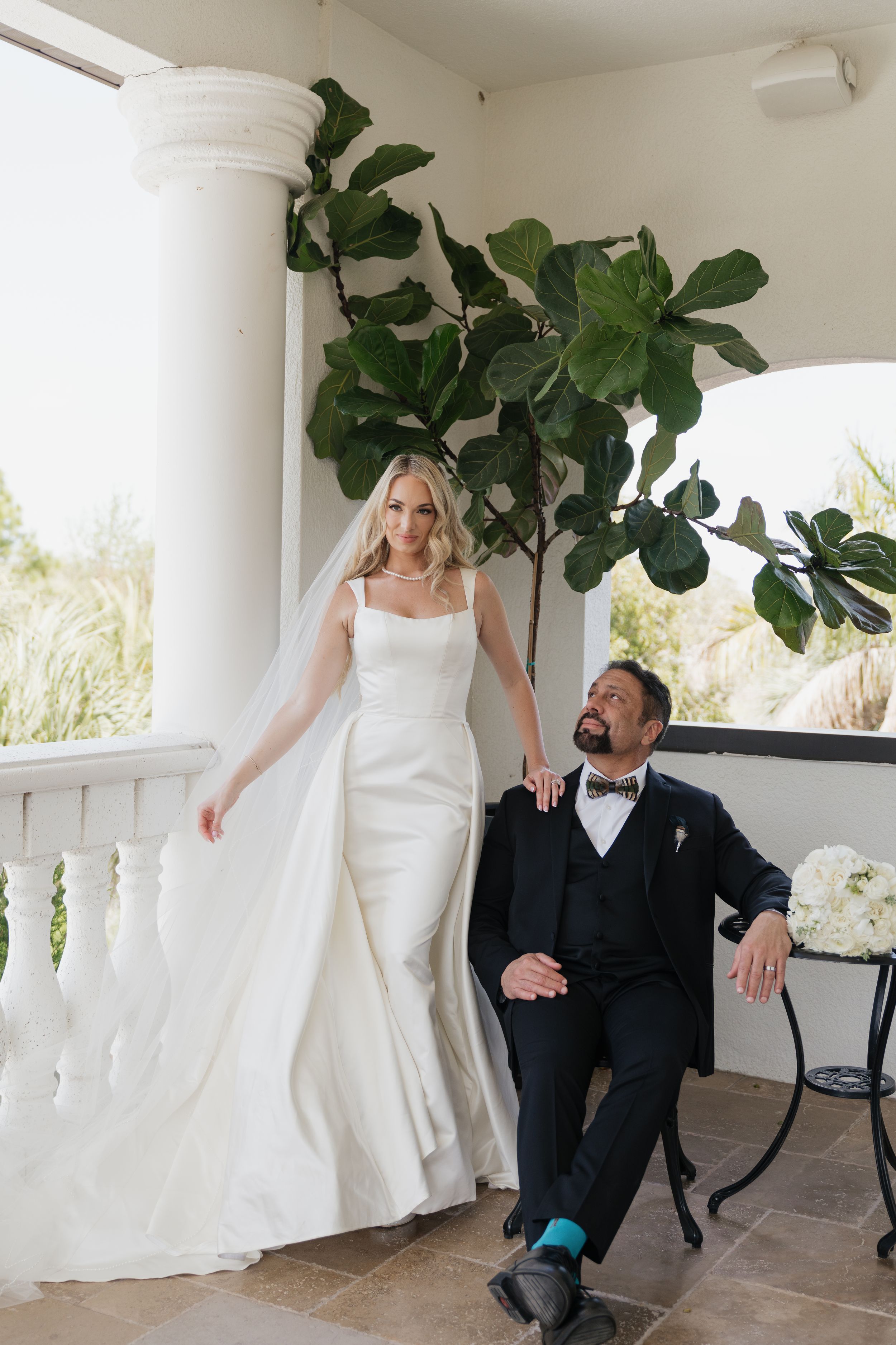 Bride in white gown and groom in navy tuxedo pose on elegant balcony with fiddle leaf fig tree backdrop.