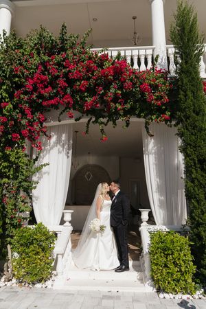 Bride and groom kissing on white colonial mansion steps adorned with vibrant red bougainvillea flowers.