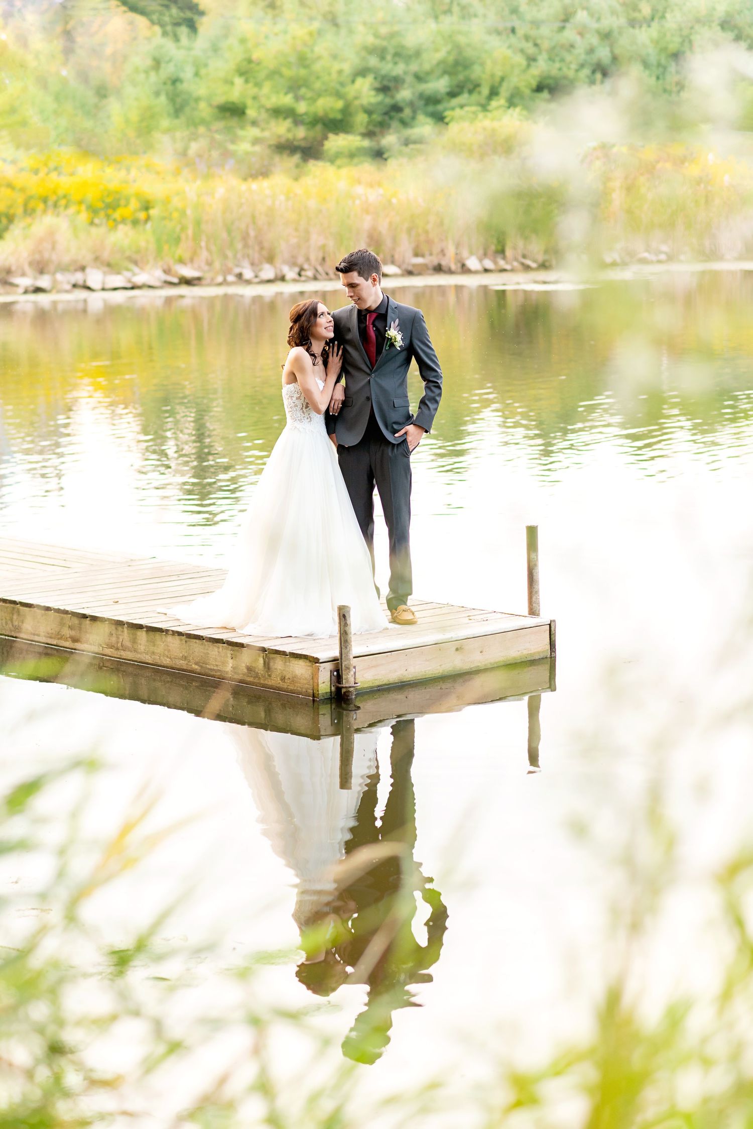 Bride and groom share a romantic moment on a wooden bridge surrounded by lush green trees in golden sunlight.